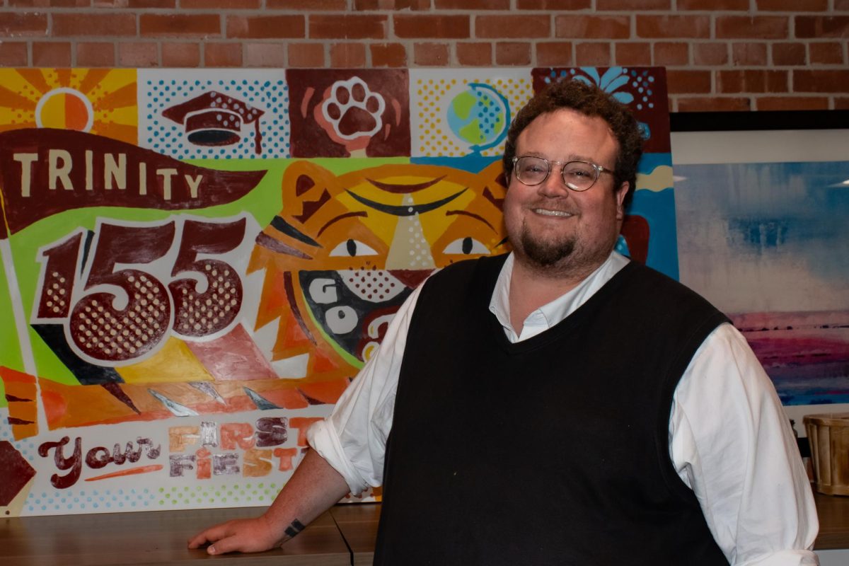 BEN WILLIAMS smiles in front of Trinity-themed artwork.